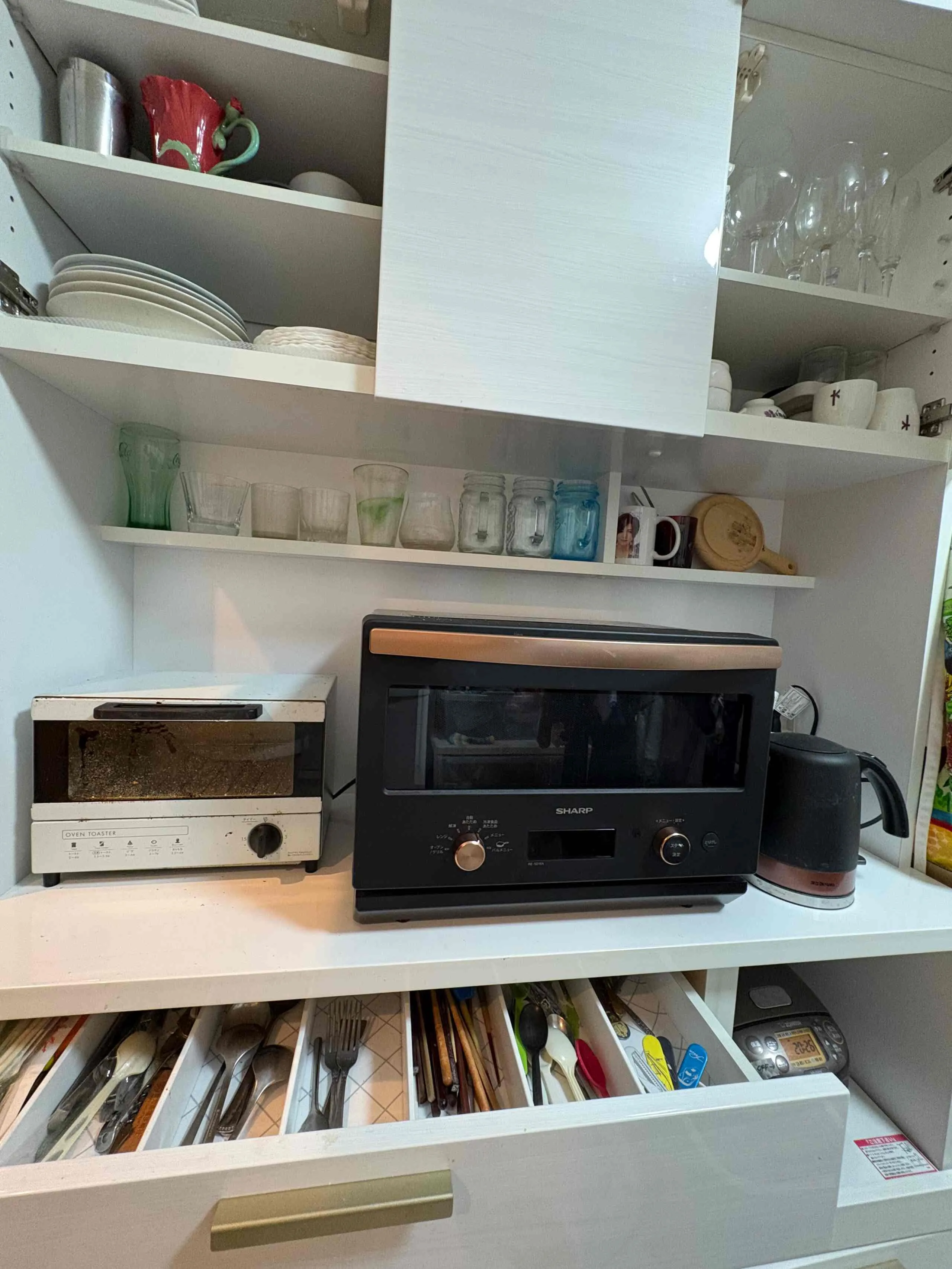 Shared kitchen shelves with plates, glasses, a microwave, kettle, and toaster oven.